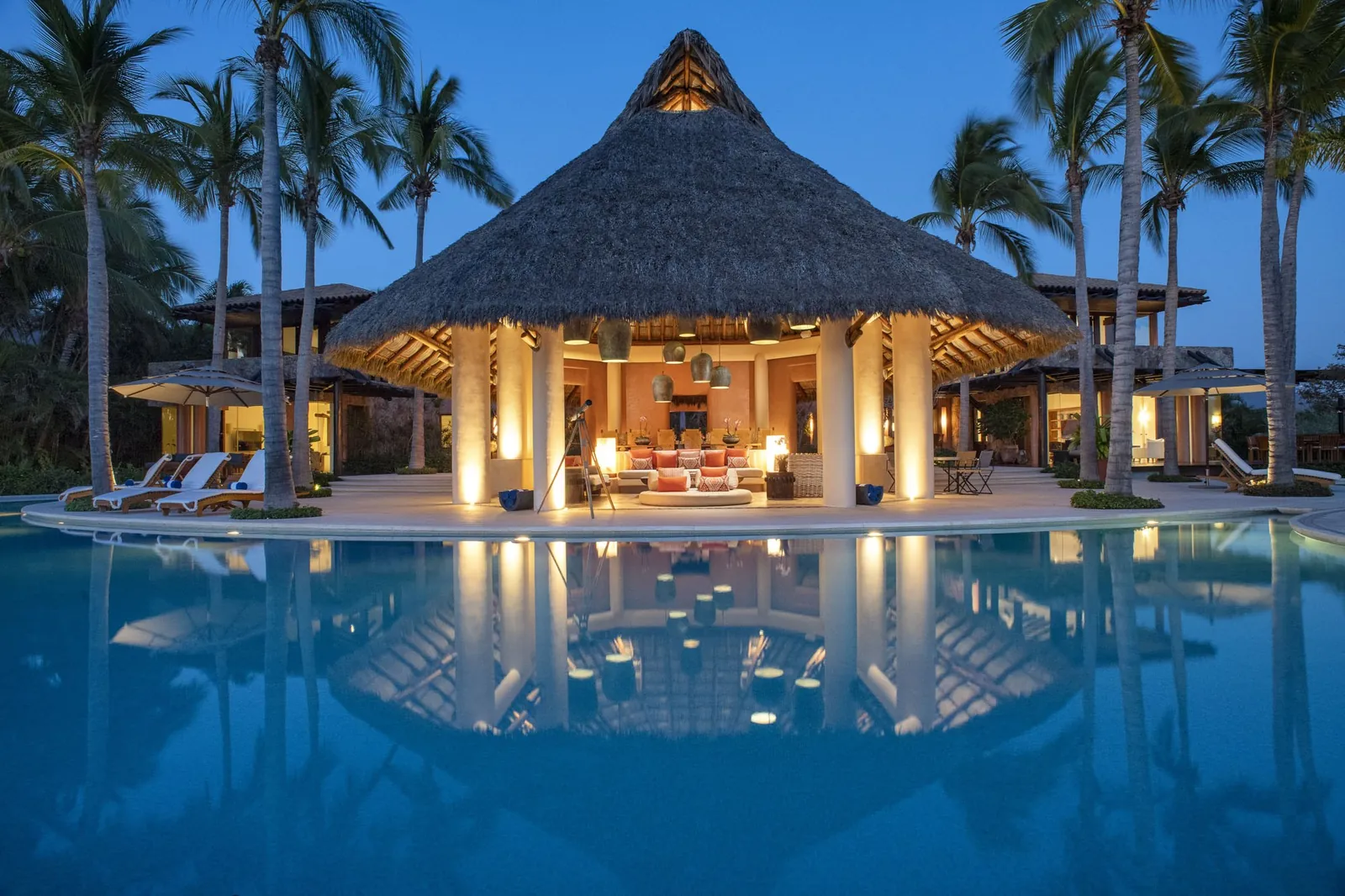 The palapa lit at dusk and reflected in the still lagoon pool
