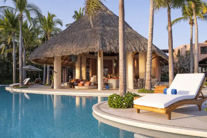 A white sun lounger at the water's edge beside the palapa